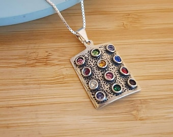 May include: A silver necklace with a rectangular pendant featuring a textured surface and 12 colorful gemstones. The pendant is decorated with a variety of colors, including red, green, blue, yellow, and purple.
