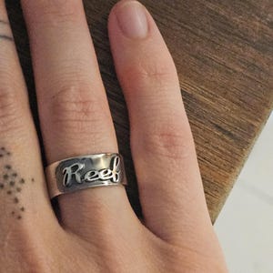 Puede incluir: Un anillo plateado con la palabra "Reef" grabada en cursiva. El anillo se lleva en un dedo, con un fondo de madera.