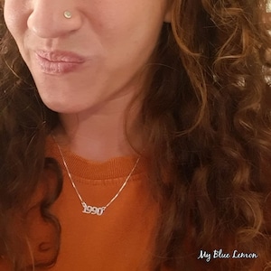 May include: A woman wearing a brown sweater with a silver necklace that says "1990". She has a small silver nose ring.