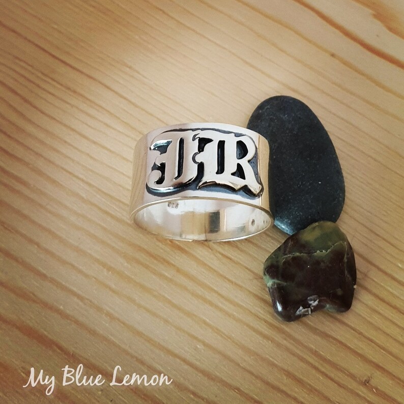 Op de afbeelding: Een zilveren ring met de letters "JFR" gegraveerd in een vet, gotisch lettertype. De ring ligt op een houten oppervlak met twee kleine stenen.