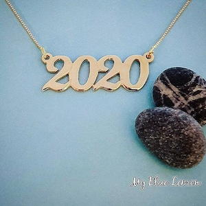 May include: A gold necklace with a 2020 pendant. The necklace is on a blue background with two gray stones.