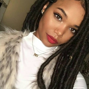 May include: A woman with dark locs and a white turtleneck. She wears a faux fur vest and a gold necklace with the number "85". Her lips are painted red, and she has a nose ring and earrings.