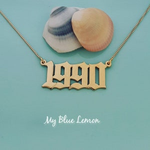 Könnte beinhalten: Goldkette mit einem 1990-Anhänger in gotischer Schrift. Die Kette liegt auf einem hellblauen Hintergrund mit zwei Muscheln.