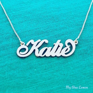 May include: A silver-toned nameplate necklace featuring the name Katie in a cursive script font. The pendant is attached to a thin box chain. The text My Blue Lemon is printed in the bottom right corner of the image.
