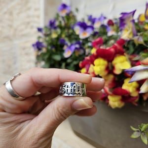 以下が含まれることがあります： ゴシック体のフォントで「Shalom」の文字が刻まれたシルバーリング。ぼやけた背景には花と窓があり、リングがその前にかざされています。