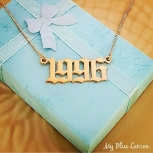 Könnte beinhalten: Goldkette mit einem 1996-Anhänger in gotischer Schrift. Die Kette liegt auf einer hellblauen Geschenkbox mit einem weißen Band.