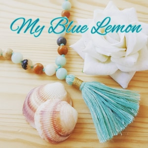 Op de afbeelding: Een turquoise blauwe kralenketting met een gouden kwast en een witte bloem. De ketting is gemaakt van verschillende stenen, waaronder turquoise, bruin en wit. De ketting ligt op een houten oppervlak met twee schelpen.
