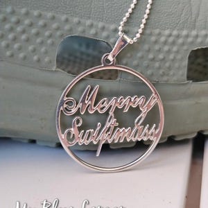 May include: Silver pendant necklace with the words "Merry Swiftmas" in a circular design. The necklace is on a silver chain. The background is a light green surface. The text "My Blue Lemon" is at the bottom.
