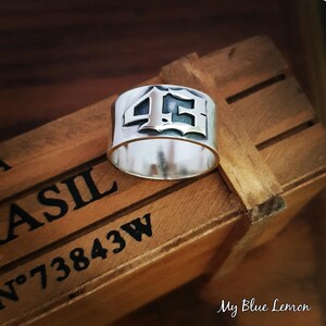 Peut inclure: Une bague en argent avec le numéro "45" gravé en police gothique. La bague est posée sur une boîte en bois avec les mots "Brasil" et "N° 73843W" imprimés dessus.