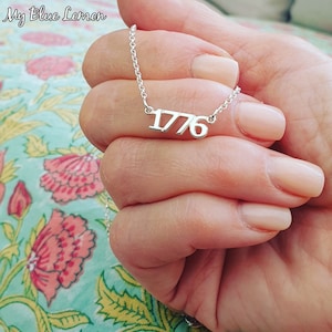 Könnte beinhalten: Eine silberne Halskette mit der Zahl 1776, die daran hängt. Die Kette wird von einer Hand mit hellrosa Nagellack gehalten.