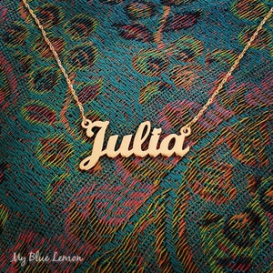 May include: Gold necklace with the name "Julia" spelled out in cursive lettering. The necklace is on a patterned fabric background.