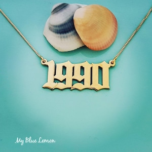 Könnte beinhalten: Goldkette mit einem 1990-Anhänger in gotischer Schrift. Die Kette liegt auf einem blauen Hintergrund mit zwei Muscheln.