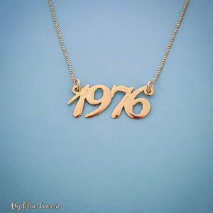 Könnte beinhalten: Eine goldene Halskette mit einem Anhänger, auf dem "1976" in einer kursiven Schrift steht.