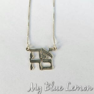 Puede incluir: Un collar con colgante plateado con letras hebreas. El colgante cuelga de una fina cadena. Las palabras "My Blue Lemon" están escritas en cursiva en la parte inferior de la imagen.