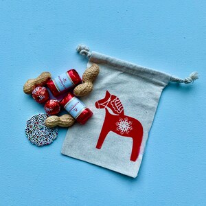Könnte beinhalten: Eine kleine weiße Stofftasche mit einem roten skandinavischen Dala-Pferd-Design. Die Tasche ist gefüllt mit rot-weiß verpackten Schokoladen und Erdnüssen.