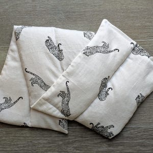 May include: Cream-coloured rectangular pillow with a black leopard print. The pillow is folded in a zig-zag pattern. The leopard print features stylized leaping leopards. The pillow is made of a soft, textured fabric.