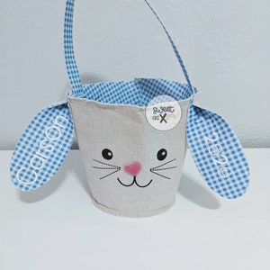 Puede incluir: Una cesta de Pascua de conejito blanca con orejas y asa de tela vichy azul y blanca. La cesta tiene una nariz rosa y ojos y bigotes negros bordados. Las orejas est&aacute;n personalizadas con los nombres "Carson" y "Zoe".