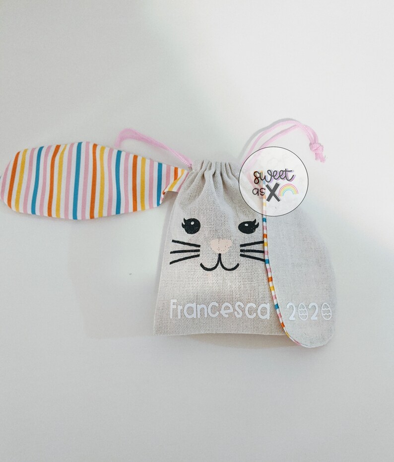Puede incluir: Una bolsa blanca de cord&oacute;n con una cara de conejo y orejas rayadas. La bolsa tiene el texto "Francesca 2020" y una peque&ntilde;a pegatina que dice "Sweet as X".