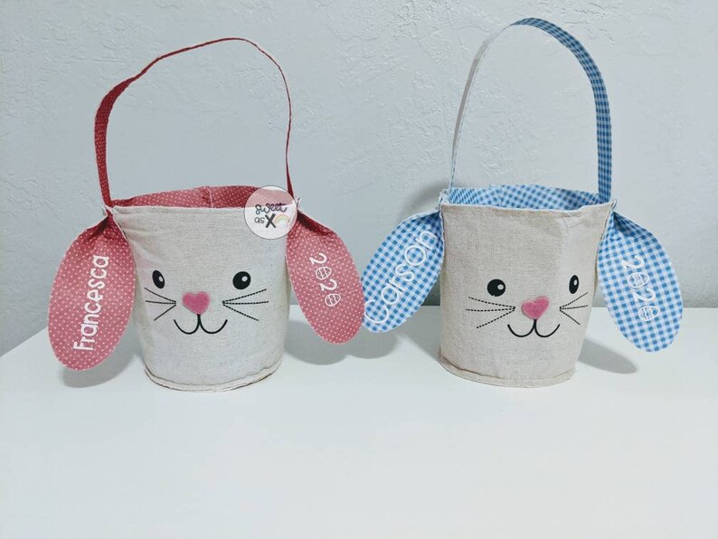 Puede incluir: Dos cestas de Pascua de conejito personalizadas, una con orejas de lunares rosas y el nombre "Francesca" y la otra con orejas de cuadros azules y el nombre "Carson".