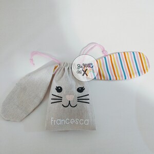 Puede incluir: Una bolsa de tela gris con forma de conejo con un cord&oacute;n de ajuste rosa y una oreja de tela a rayas. La bolsa tiene una cara de conejo en blanco y negro con el nombre "Francesca" impreso en la parte delantera.
