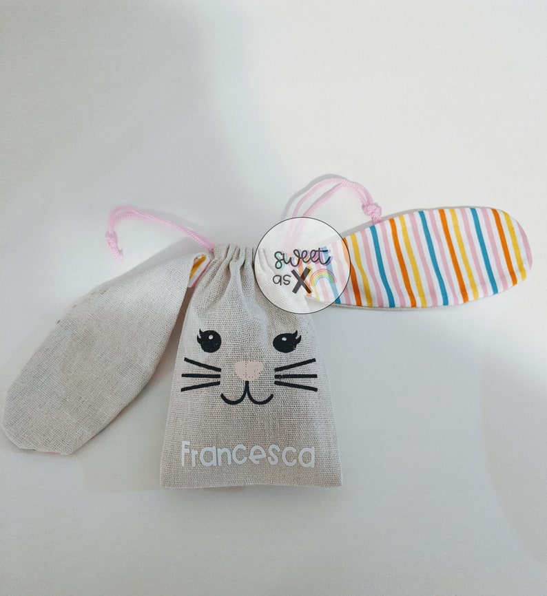 Puede incluir: Una bolsa de tela gris con forma de conejo con un cord&oacute;n de ajuste rosa y una oreja de tela a rayas. La bolsa tiene una cara de conejo en blanco y negro con el nombre "Francesca" impreso en la parte delantera.