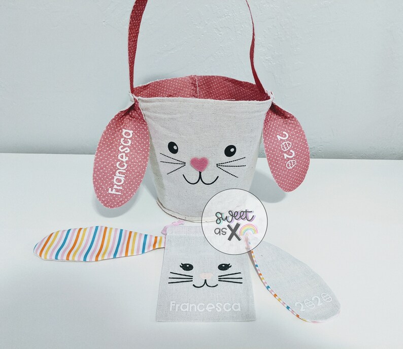 Puede incluir: Una cesta de Pascua de conejito blanca con orejas rosas y un asa rosa. Las orejas est&aacute;n personalizadas con el nombre "Francesca" y el a&ntilde;o "2023".