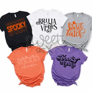 May include: A collection of Halloween-themed t-shirts in various colors. The shirts feature spooky phrases and graphics, including "Spooky," "Bruja Vibes," "Basic Witch," "Hello Pumpkin," and "Spooky Mama." The designs are in orange and black.