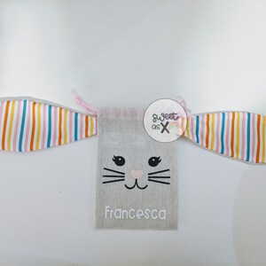 Puede incluir: Una bolsa de cord&oacute;n de color gris claro con forma de conejo, con orejas rosas, una cara bordada en negro y el nombre "Francesca" escrito debajo. La bolsa tiene un patr&oacute;n de rayas de arco&iacute;ris en las orejas.