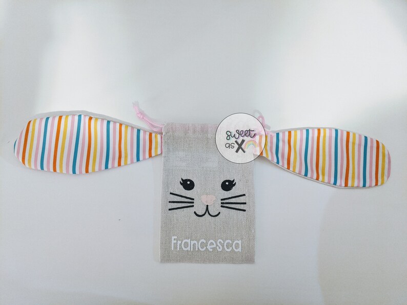 Puede incluir: Una bolsa de cord&oacute;n de color gris claro con forma de conejo, con orejas rosas, una cara bordada en negro y el nombre "Francesca" escrito debajo. La bolsa tiene un patr&oacute;n de rayas de arco&iacute;ris en las orejas.
