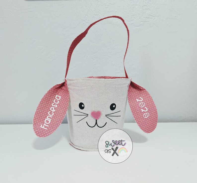 Puede incluir: Una cesta de Pascua blanca con forma de conejo con orejas de lunares rosas. Las orejas est&aacute;n personalizadas con el nombre "Francesca" y el a&ntilde;o "2022".