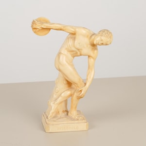 May include: A cream-colored sculpture of a discus thrower in a dynamic athletic pose, standing on a rectangular base. The figure is depicted in a forward-leaning stance with one arm extended holding a disc and the other reaching toward the leg.