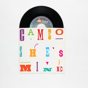 May include: A black vinyl record partially inserted into a white sleeve. The sleeve features the text "CAMEO SHE'S MINE" in various bright colors. The record label is visible, with the text "ATLANTA ARTISTS".
