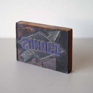 May include: A wooden printing block with a blue and silver design. The design features two arrows pointing towards each other and the word "SOLIOSO" in the centre.