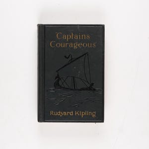 May include: A black hardcover book titled "Captains Courageous" by Rudyard Kipling. The cover features a gold embossed title and a detailed illustration of a sailboat. The book is set against a white background.