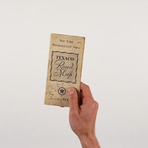 以下が含まれることがあります： ニューヨーク都市圏のビンテージテキサコロードマップ。白い背景に、手に持った地図。地図には星のロゴと「Tour with TEX」の文字があります。