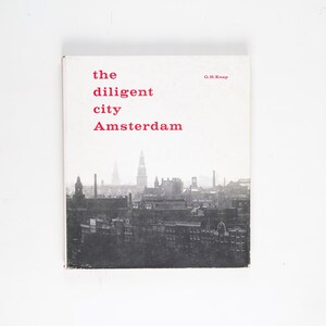 May include: A book cover with the title "The Diligent City Amsterdam" in red text. The cover features a black and white photograph of the Amsterdam skyline.