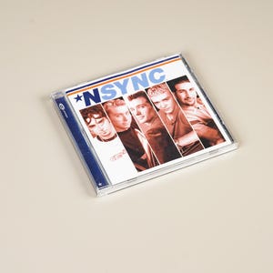 May include: A compact disc case featuring the band *NSYNC. The album cover displays six men in a collage of headshots. The band's name is printed in blue and white, with a star graphic. The case is clear with a blue spine.
