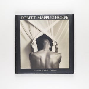 May include: A monochrome book cover with the title "ROBERT MAPPLETHORPE" at the top. The image depicts a person from behind, holding fabric. The text "Foreword by Ntozake Shange" is at the bottom of the cover.