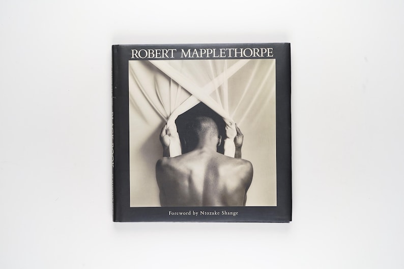 May include: A monochrome book cover with the title "ROBERT MAPPLETHORPE" at the top. The image depicts a person from behind, holding fabric. The text "Foreword by Ntozake Shange" is at the bottom of the cover.