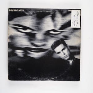 Puede incluir: Portada de álbum en blanco y negro con una imagen borrosa de una cara y la cara de un hombre en la esquina inferior derecha. El título del álbum es "Thrashing Doves" y el texto "Feels on the Fly" está escrito en una pegatina blanca.