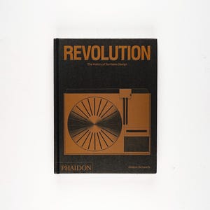 May include: A hardcover book titled "REVOLUTION" with the subtitle "The History of Turntable Design." The cover is black with gold lettering and a gold illustration of a turntable. The book is published by Phaidon.