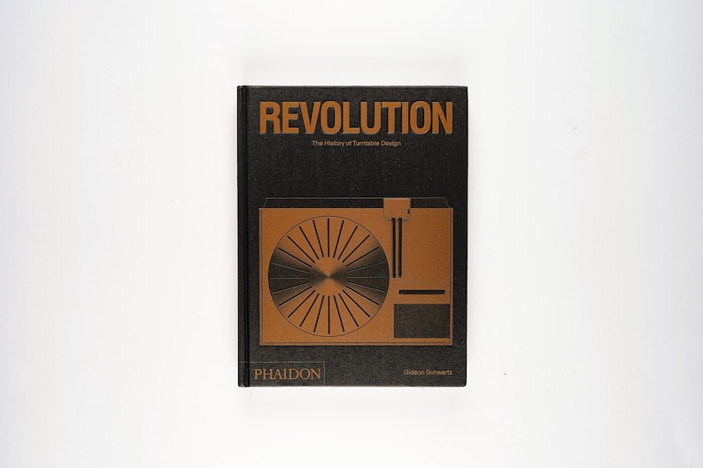 May include: A hardcover book titled "REVOLUTION" with the subtitle "The History of Turntable Design." The cover is black with gold lettering and a gold illustration of a turntable. The book is published by Phaidon.