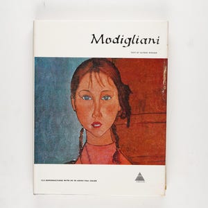 May include: A book cover featuring a portrait of a young woman with brown hair in braids, blue eyes, and a pink top. The title "Modigliani" is at the top, with text below the portrait. The cover has a white background.