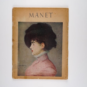 May include: A vintage book cover featuring a portrait of a woman wearing a black hat and a pink dress. The title "MANET" is printed in large letters at the top of the cover.