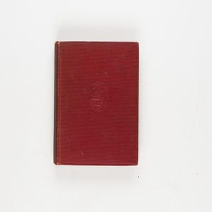 May include: A vintage, hardcover book with a deep red cover. The book has a slightly worn appearance, suggesting age. A small, embossed design is visible in the center of the cover. The book is rectangular and appears to be closed.