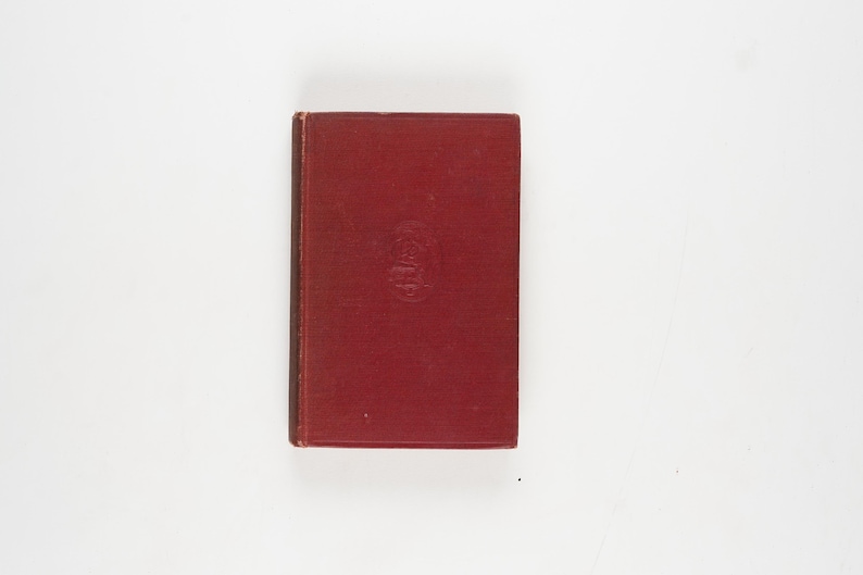 May include: A vintage, hardcover book with a deep red cover. The book has a slightly worn appearance, suggesting age. A small, embossed design is visible in the center of the cover. The book is rectangular and appears to be closed.