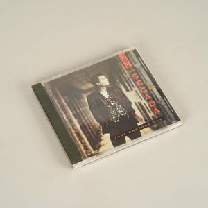 May include: A CD case featuring a man in a black jacket and patterned shirt. The CD case has the text "Just Another Day" and the artist's name "J. Secada" in red and white lettering.