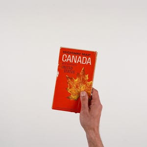 Peut inclure: Une carte routière vintage du Canada et du nord des États-Unis. La couverture est orange vif avec le mot "CANADA" en lettres blanches en gras. Une illustration détaillée d'une feuille d'érable est présentée sur la couverture.