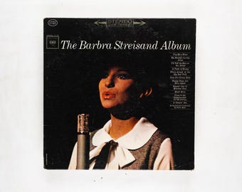 May include: Vintage vinyl record album cover for "The Barbra Streisand Album." The cover features a black background with a portrait of Barbra Streisand and a list of song titles. A microphone is in front of her.