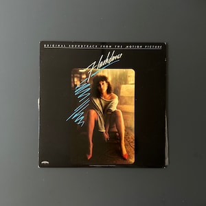 May include: A black vinyl record album cover with the title "Flashdance" in blue and white lettering. The cover features a photo of a woman in a grey sweater and red shoes sitting on a chair.